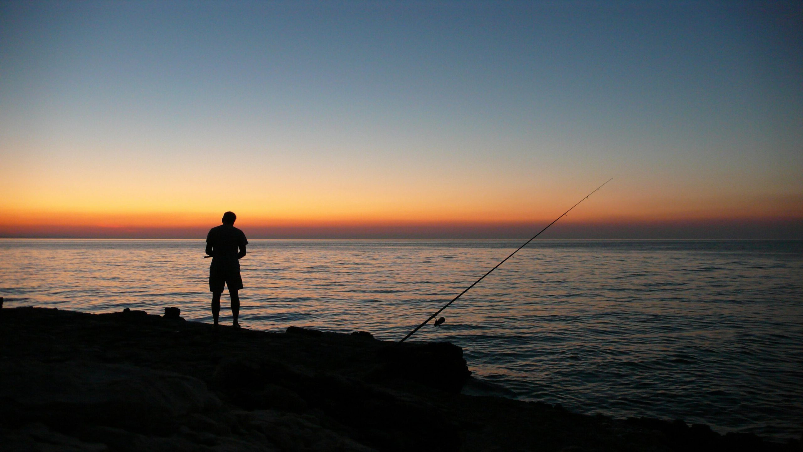 A man silhouetted against a sunset sky, fishing by the ocean. Serene and tranquil seascape.